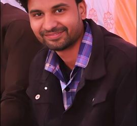 Mohammad Farhan, a man with short, curly black hair and a beard, is smiling while sitting indoors. He is wearing a black jacket over a blue plaid shirt, with a patterned backdrop featuring floral designs behind him.